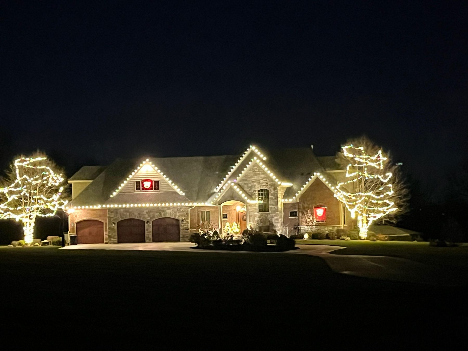 Large stone Indiana home with warm white Christmas lights along the rooflines, two mature trees wrapped trunk to branch and red lighted wreaths on the gables
