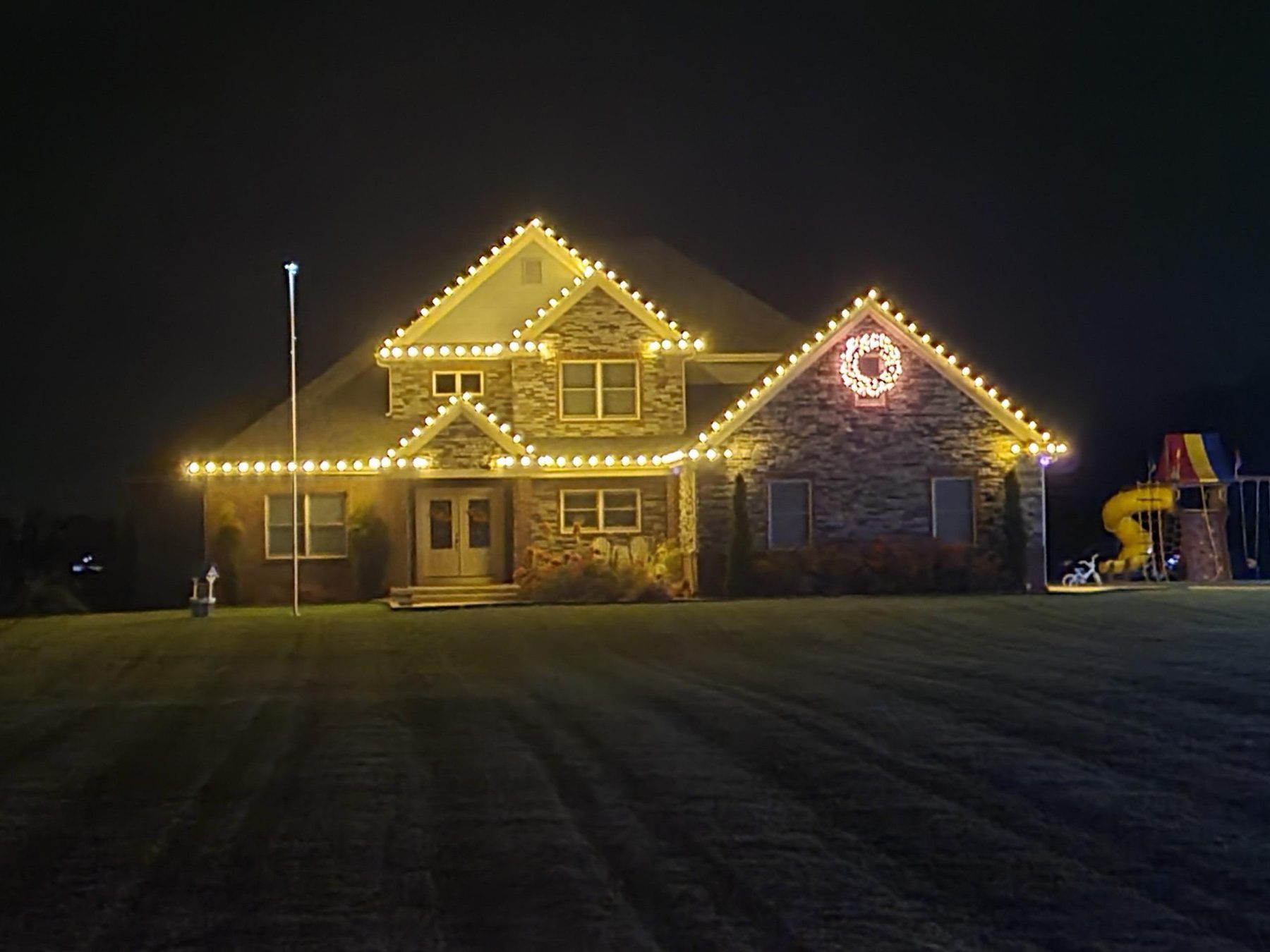 Stone Indiana home with warm white Christmas lights along every gable and eave and a lighted wreath on the right gable